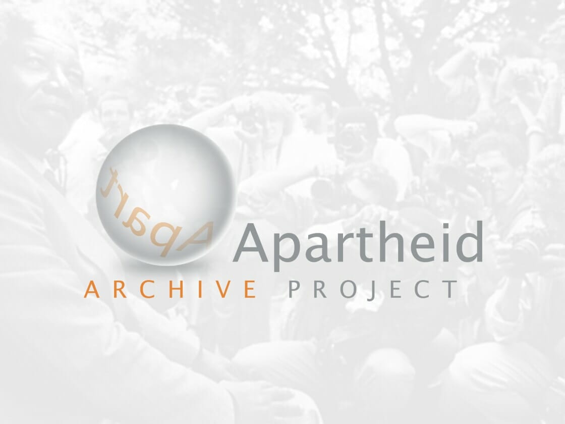 A semi-transparent logo that reads "Apartheid Archive Project" overlays a black-and-white photograph of a historic event, with a large group of photographers surrounding a seated individual in the background. The word "Aparheid" appears mirrored inside a reflective sphere on the left side of the logo.