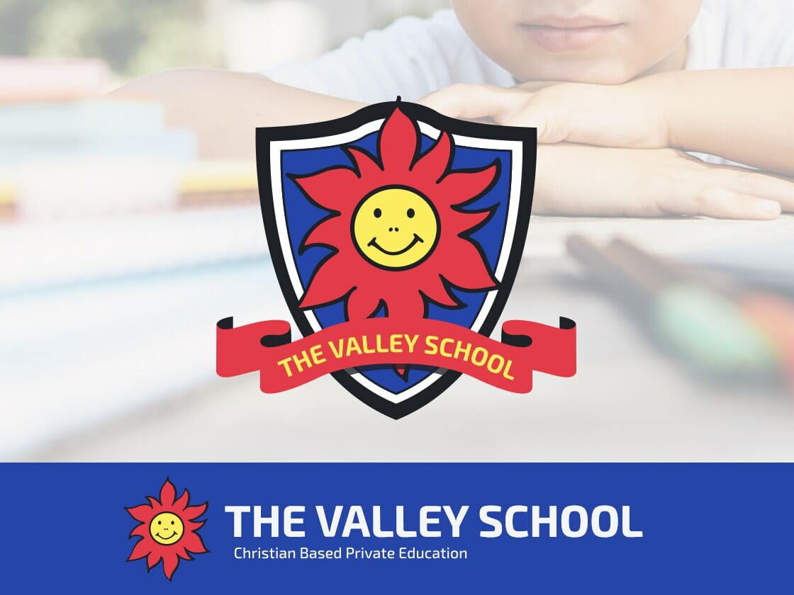 A child leans over a desk, partially shown in the background. In the foreground, there is a shield-shaped emblem with a smiling sun and the text "THE VALLEY SCHOOL" on a red banner. Below the emblem, "THE VALLEY SCHOOL" and "Christian Based Private Education" are written on a blue background.