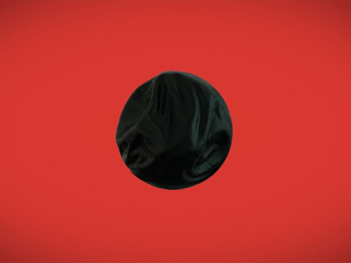 A black balloon against a vibrant red background. The balloon appears slightly deflated and wrinkled, creating irregular folds and creases on its surface. The bright background contrasts sharply with the dark color of the balloon, reminiscent of a 2018 teaser video emphasizing its shape and texture.